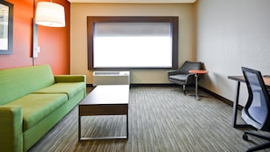 Habitación de hotel con un sofá verde, una mesa pequeña, un sillón gris, una mesita de noche roja y un ventanal grande con persianas.