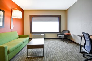 A hotel room with a green sofa, a small table, a grey armchair, a red side table, and a large window with blinds.