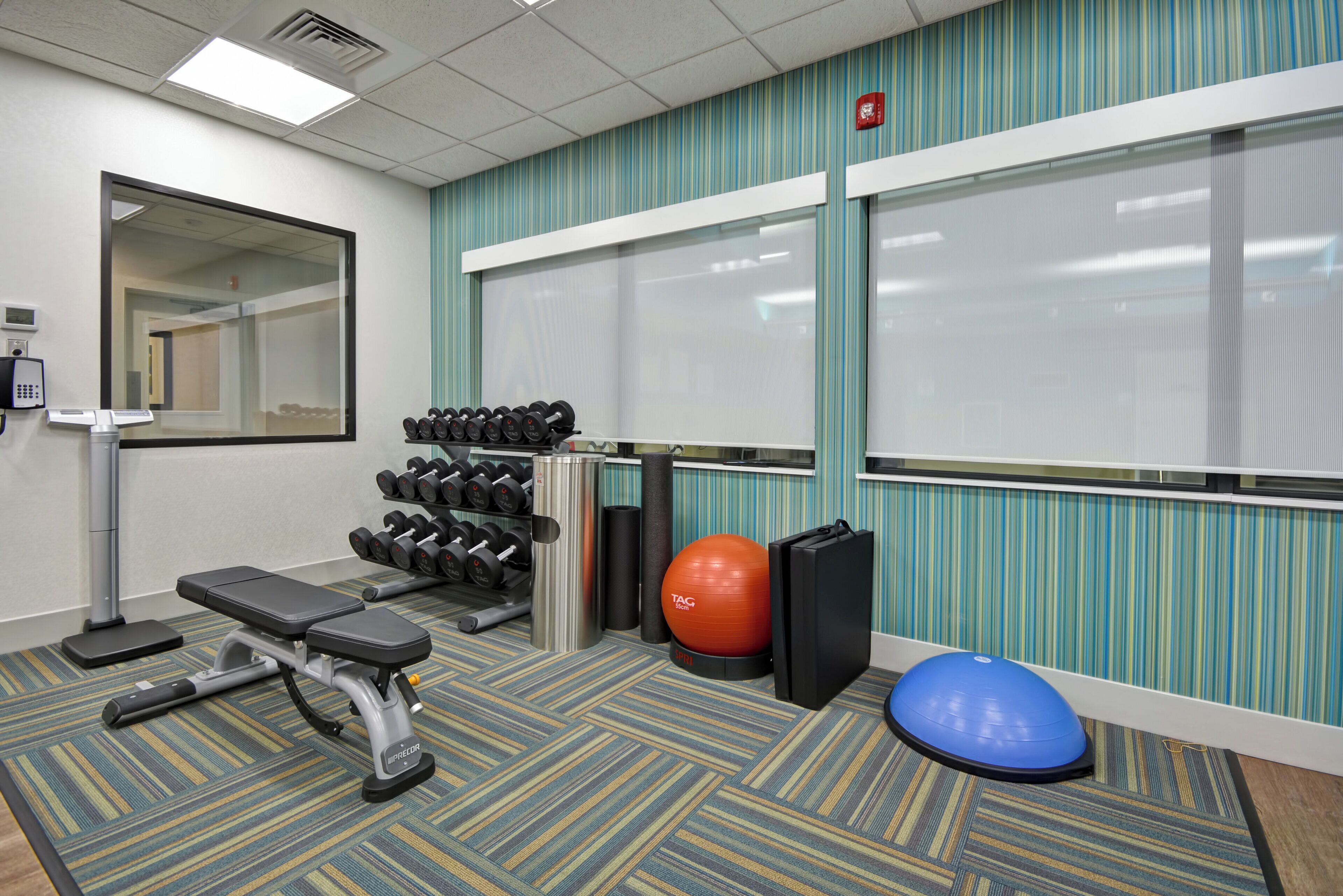 A gym with exercise equipment, a bench, a wall-mounted mirror, and a window with blinds.