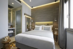 A modern hotel room with a large bed, a bedside table, a nightstand, a mirror, and a bathroom with a shower and a toilet. - Fileria Suites (Santorini)