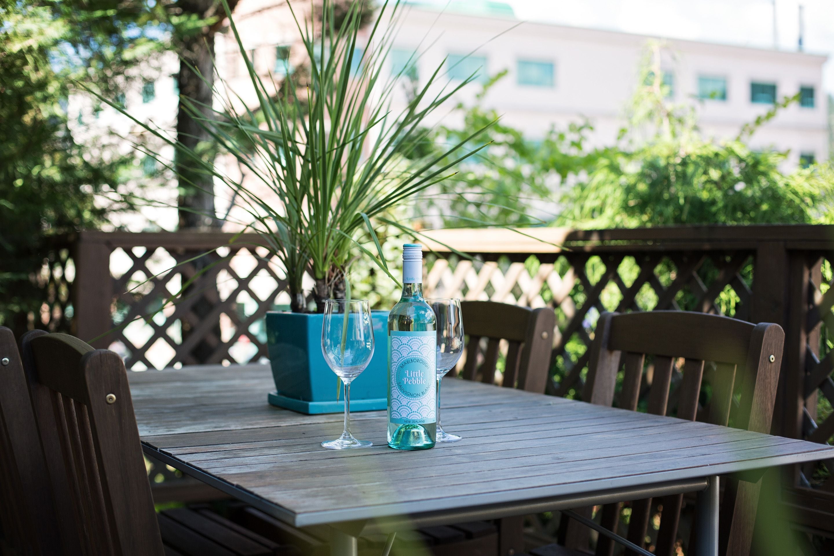 Una mesa de madera con una botella de Little Pebble y dos vasos, rodeada de sillas, en un patio exterior con valla de madera y follaje al fondo.