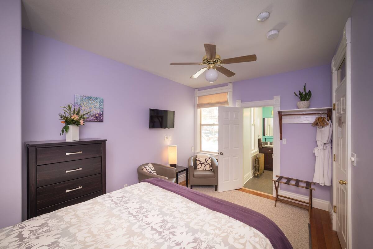 A bedroom with a bed, a dresser, a chair, a ceiling fan, a TV, and a bathroom visible through an open door.