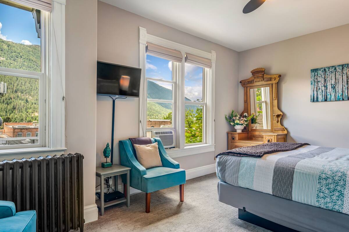 A bedroom with a bed, a chair, a TV, and a large window with a view of greenery and mountains.