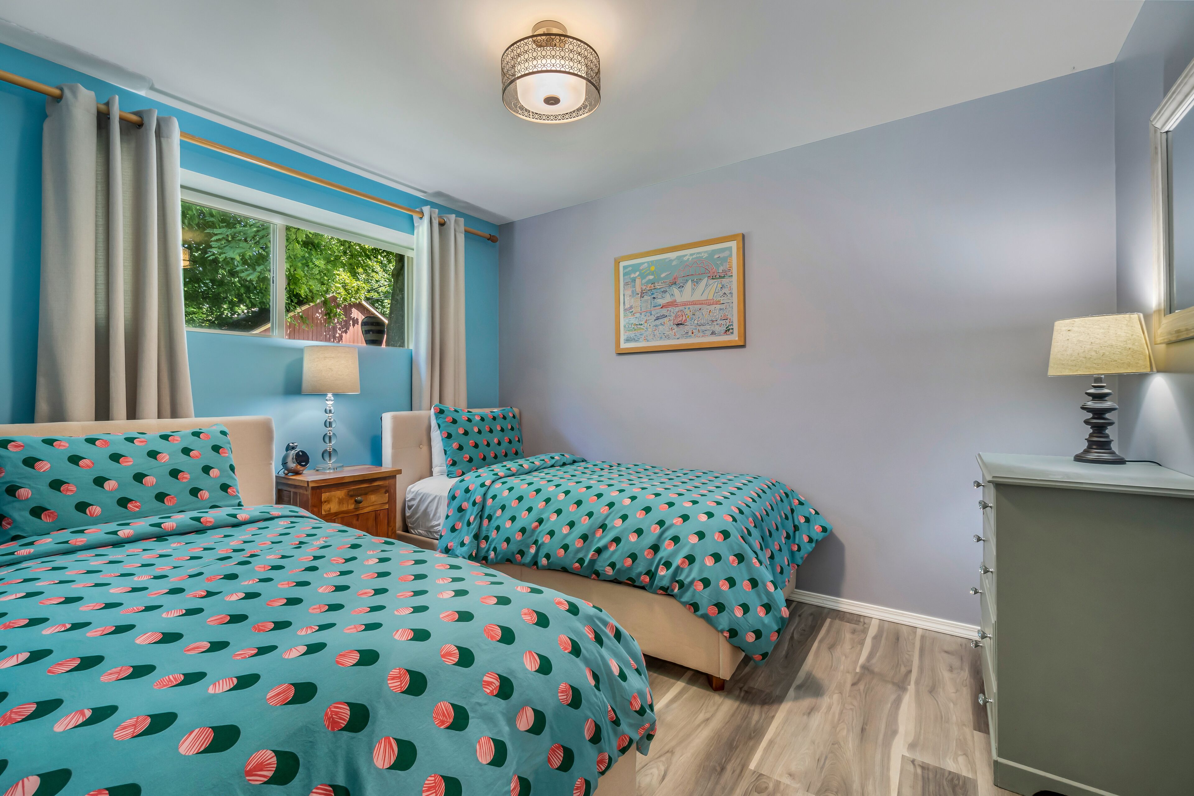 A bedroom with two beds, a nightstand, a dresser, a window with curtains, and a wall-mounted lamp.