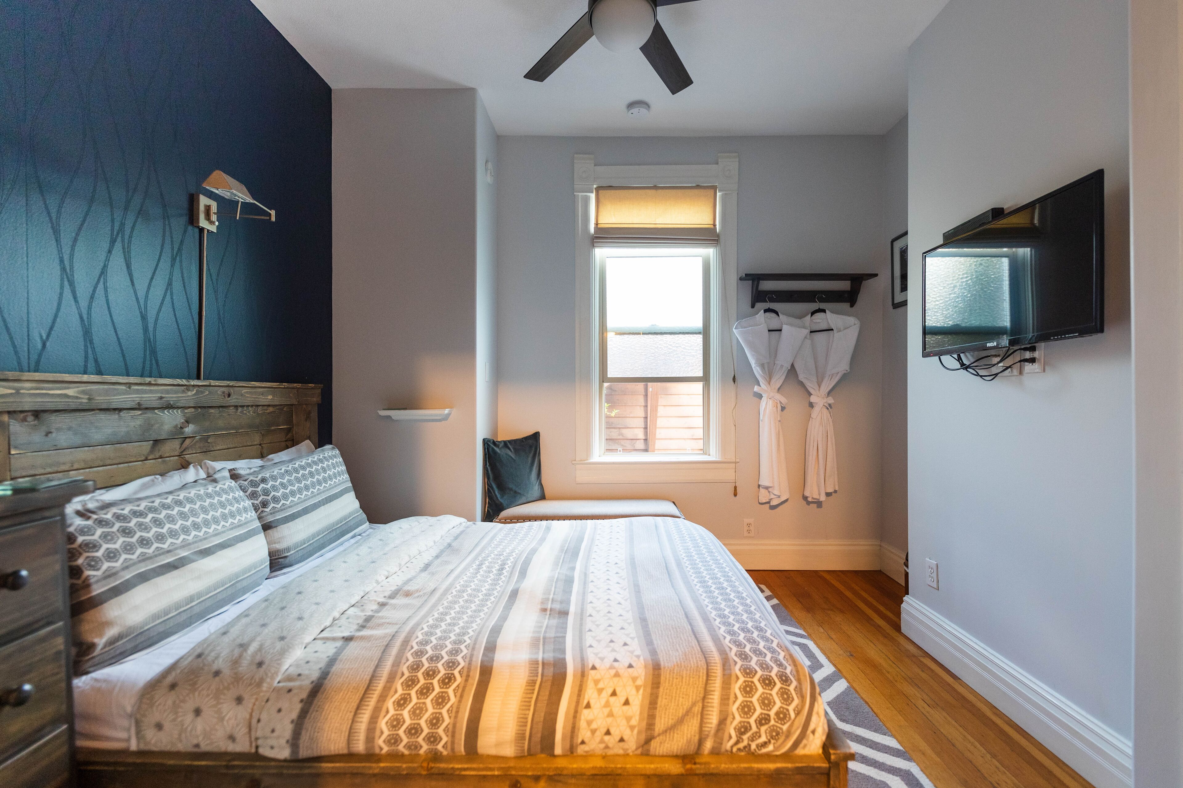 A bedroom with a bed, a ceiling fan, a window with curtains, a TV mounted on the wall, and a wooden floor.