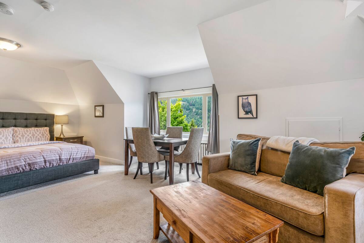 A bedroom with a bed, a wooden bench, a sofa, and a dining area with chairs and a table.