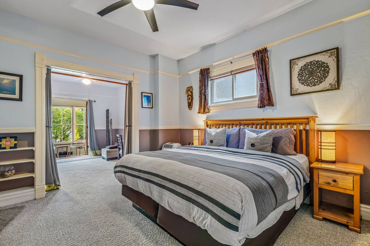 A bedroom with a large bed, a wooden nightstand, a ceiling fan, a window with curtains, and a view of the outdoors.