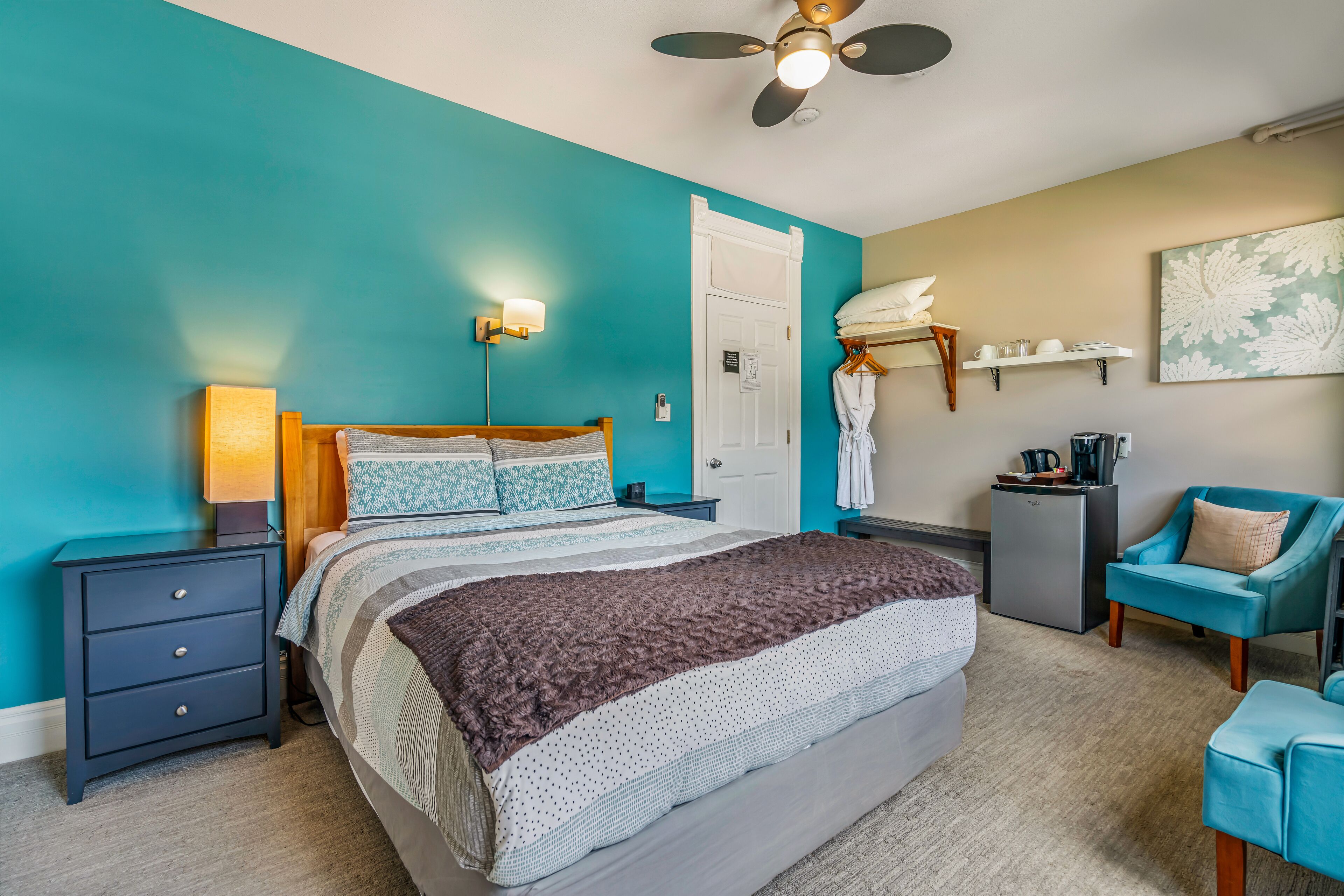 Ein Schlafzimmer mit türkisfarbenen Wänden, einem Bett mit gemustertem Überwurf, einem Nachttisch mit Lampe, einem Deckenventilator, einem kleinen Kühlschrank, einem Sessel und einem an der Wand befestigten Regal mit Handtüchern.