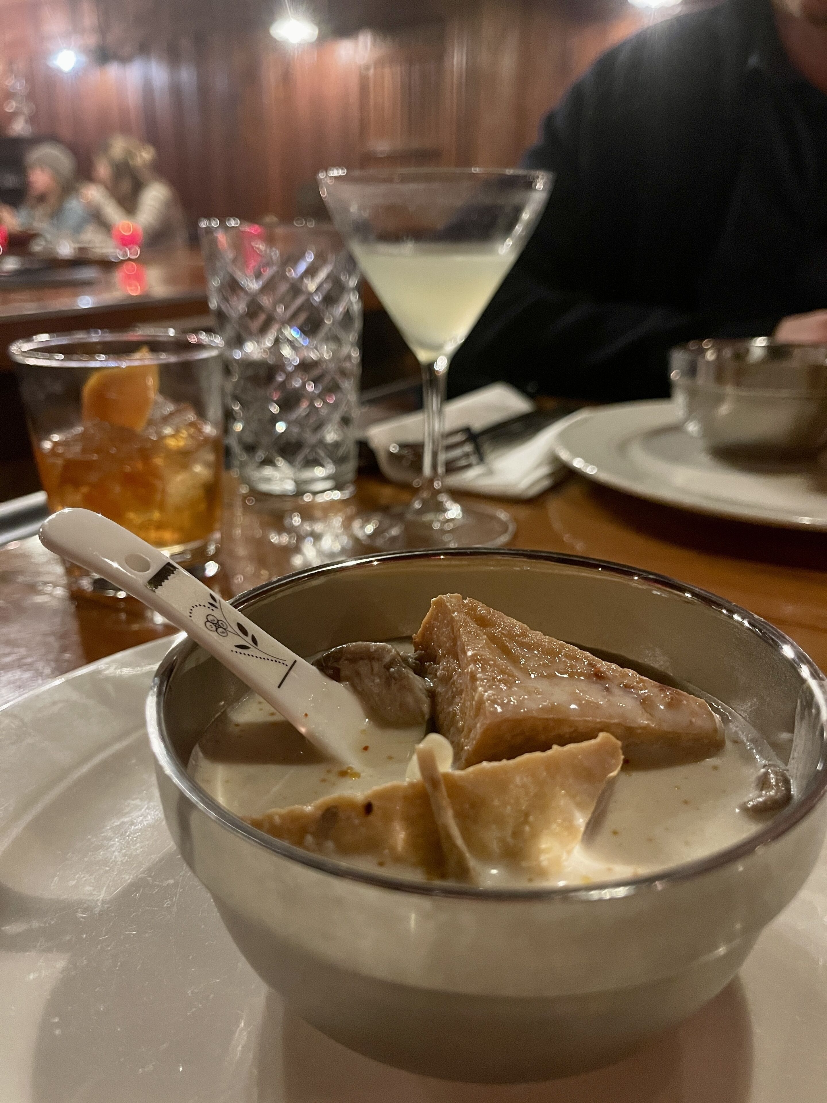 A dining table with a bowl of soup and a spoon, a glass of iced tea, and a martini glass.