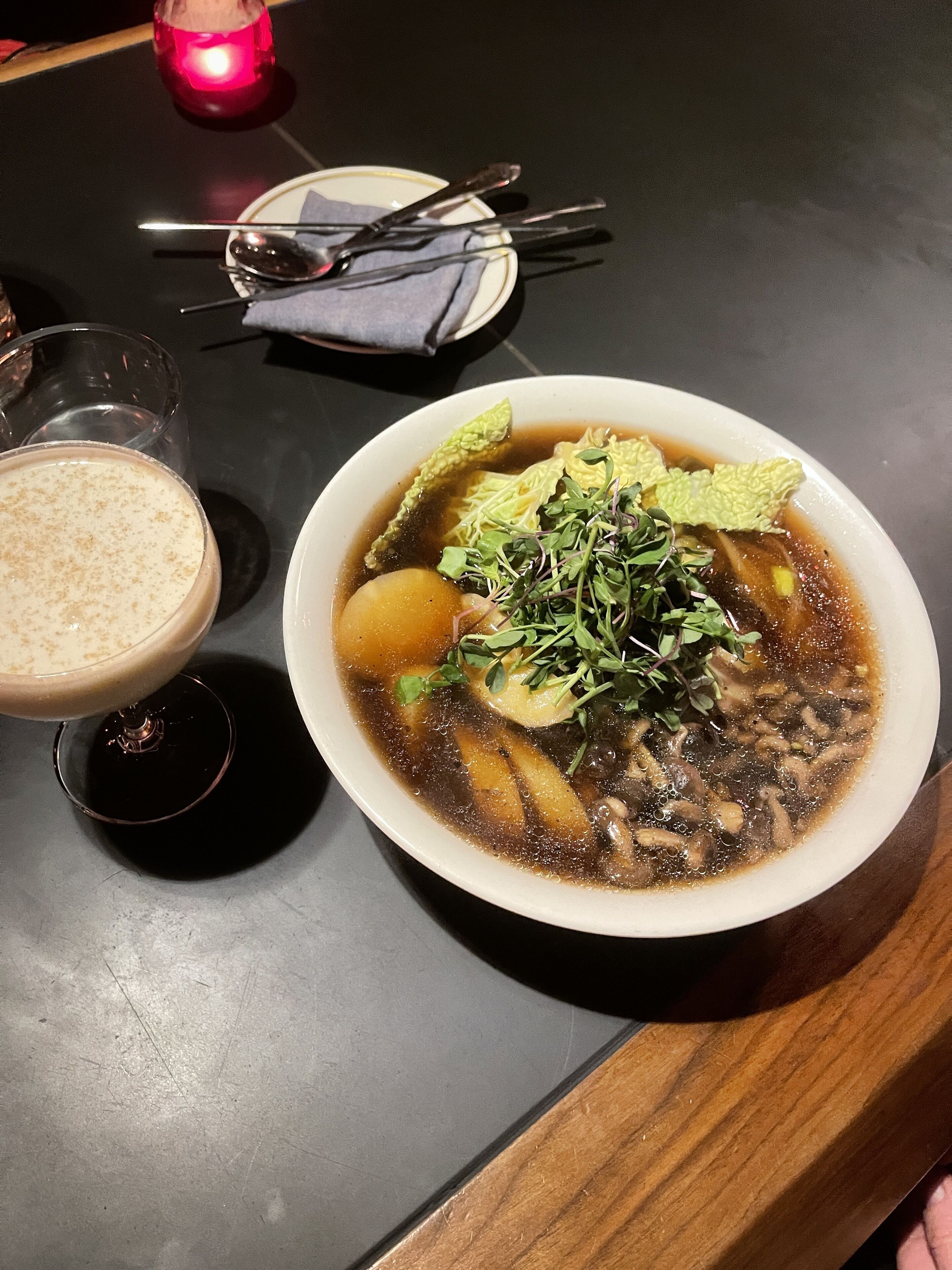 A bowl of soup with various ingredients, a glass of a creamy beverage, and a candle on a table.