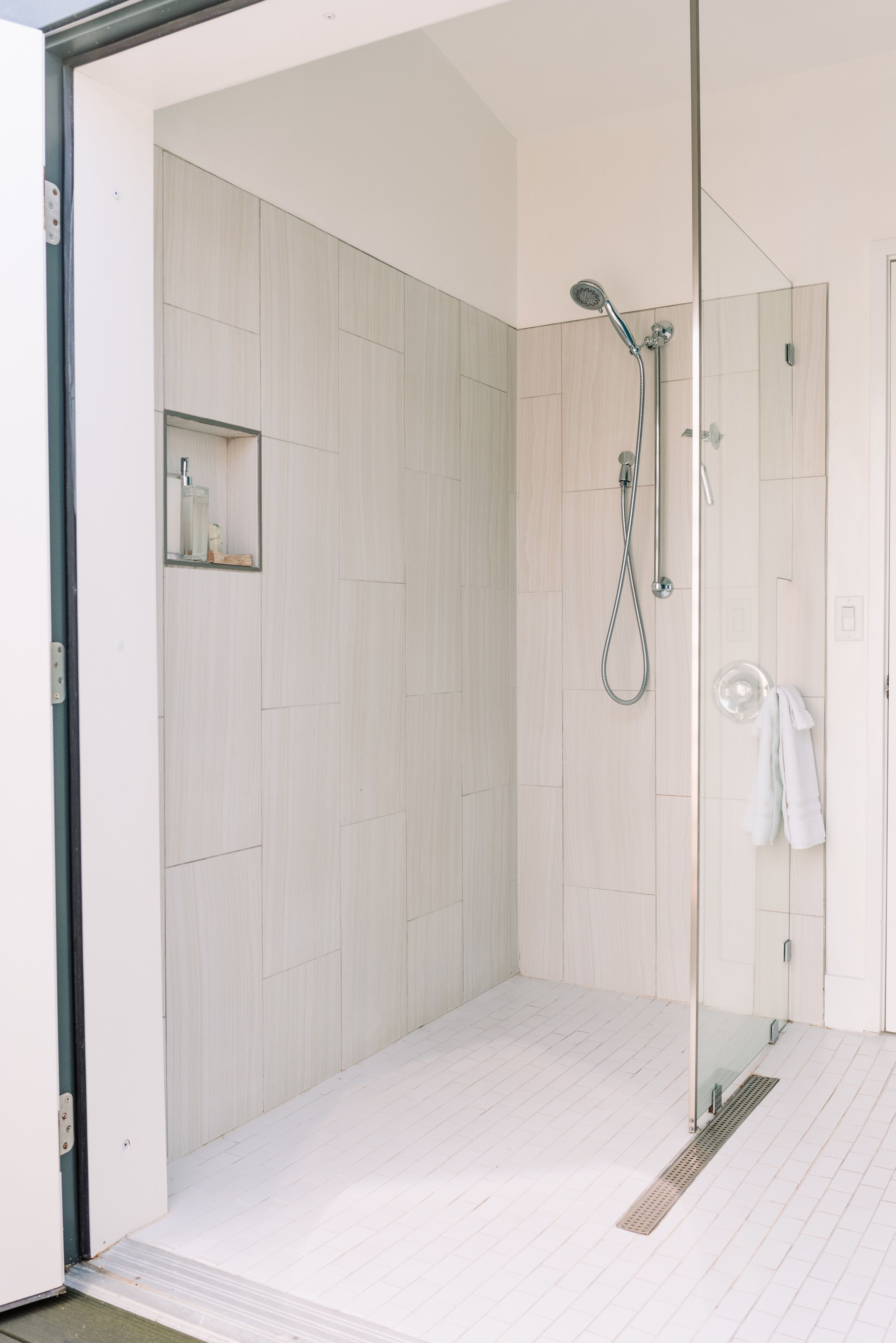 Une salle de bain moderne avec une douche à parois de verre, un carrelage blanc au sol et un petit miroir agrémenté d’accessoires de toilette.