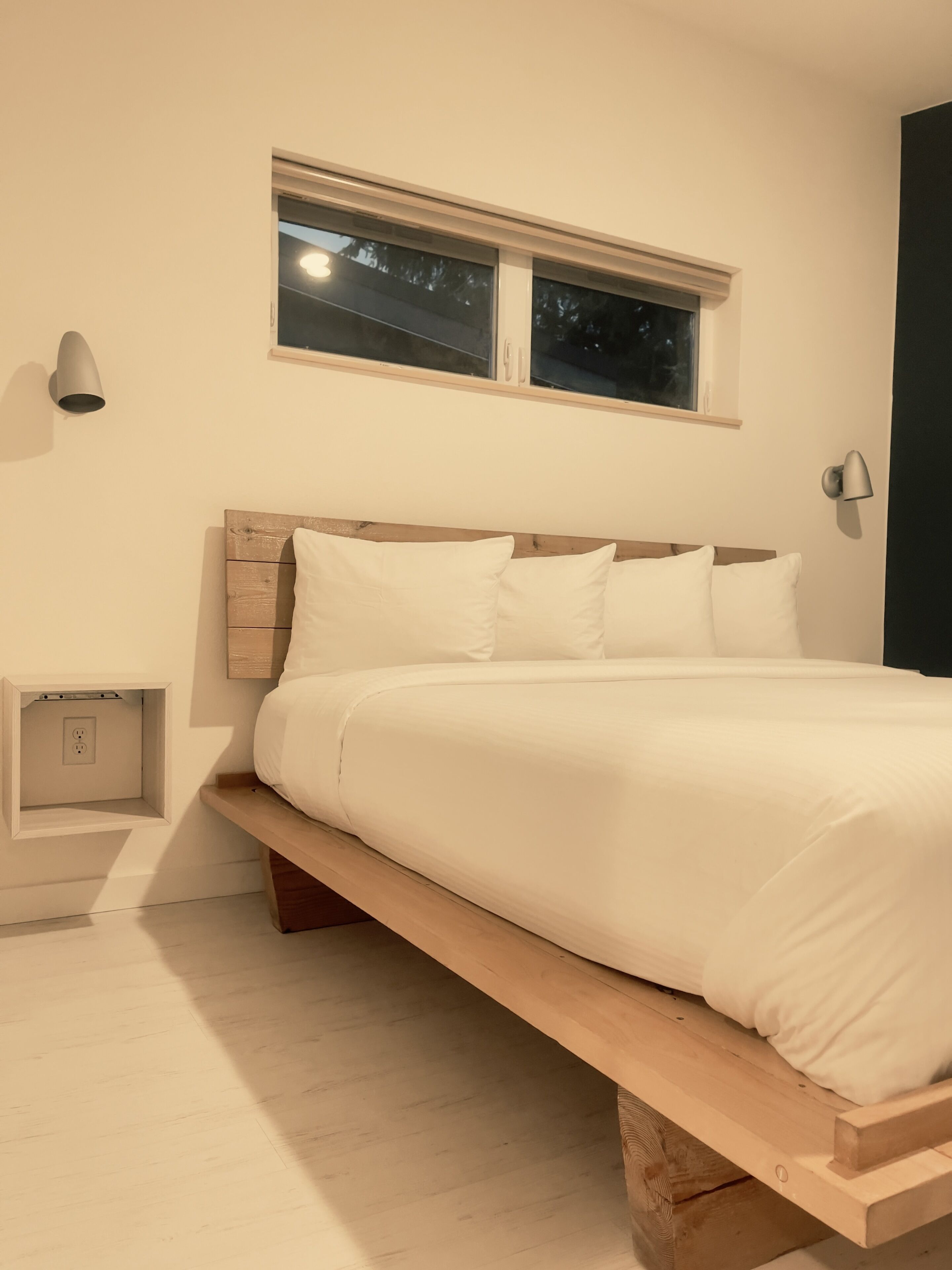 A bedroom with a wooden bed frame, white bedding, a nightstand, a wall-mounted lamp, and a window with a view of trees.