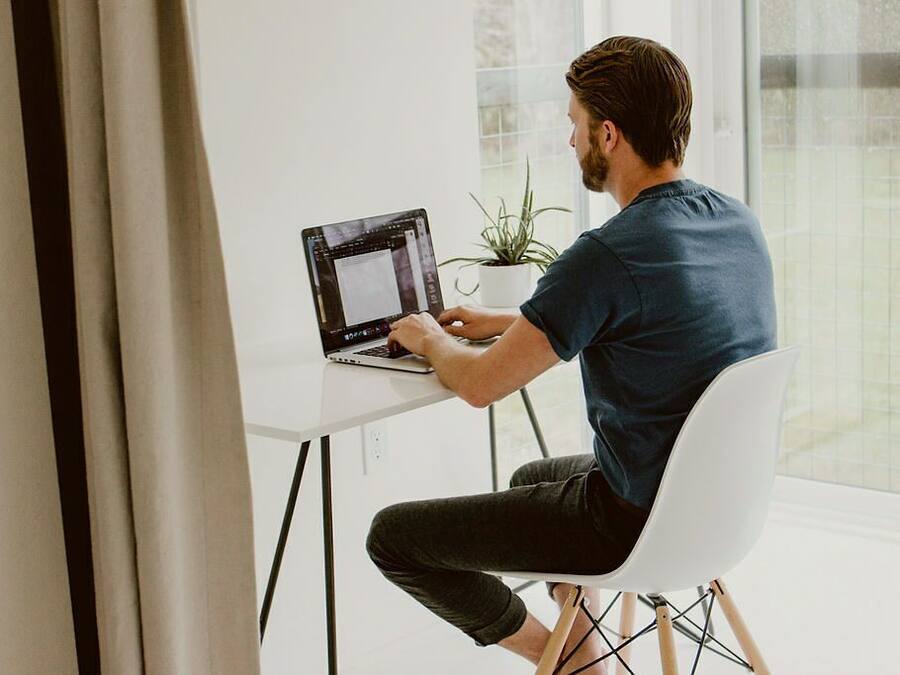 A person sitting at a white desk with a laptop, a potted plant, and a window in the background.
