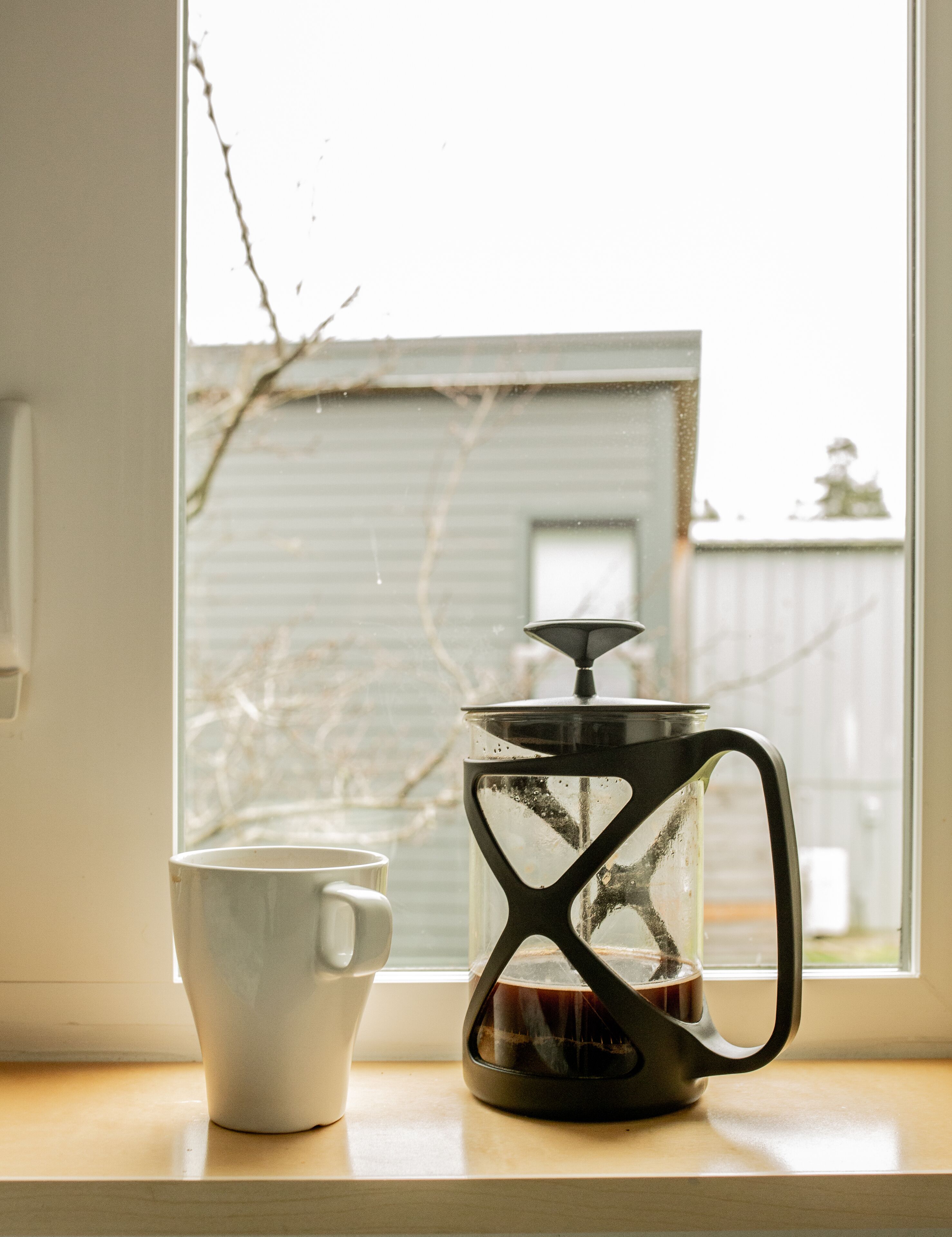 A coffee maker and a mug on a windowsill with a view of a house.