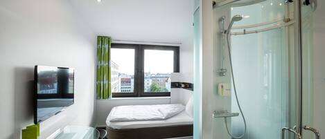 Une chambre d’hôtel moderne équipée d’un lit, d’un bureau en verre, d’une télévision, d’une fenêtre avec des rideaux et d’une cabine de douche en verre.