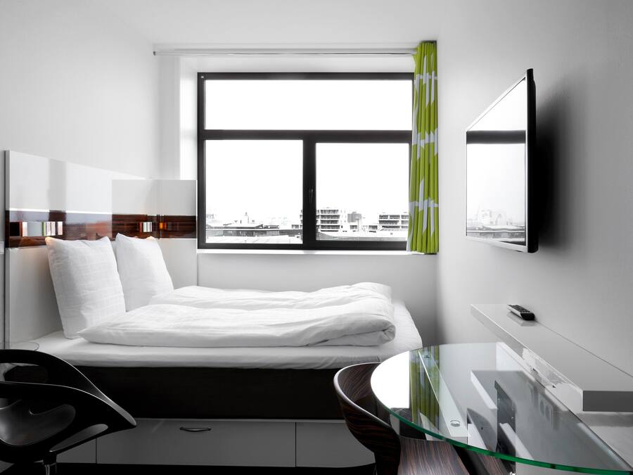 A modern hotel room with a bed, a desk with a glass top, a chair, a TV mounted on the wall, and a window with a view of buildings.