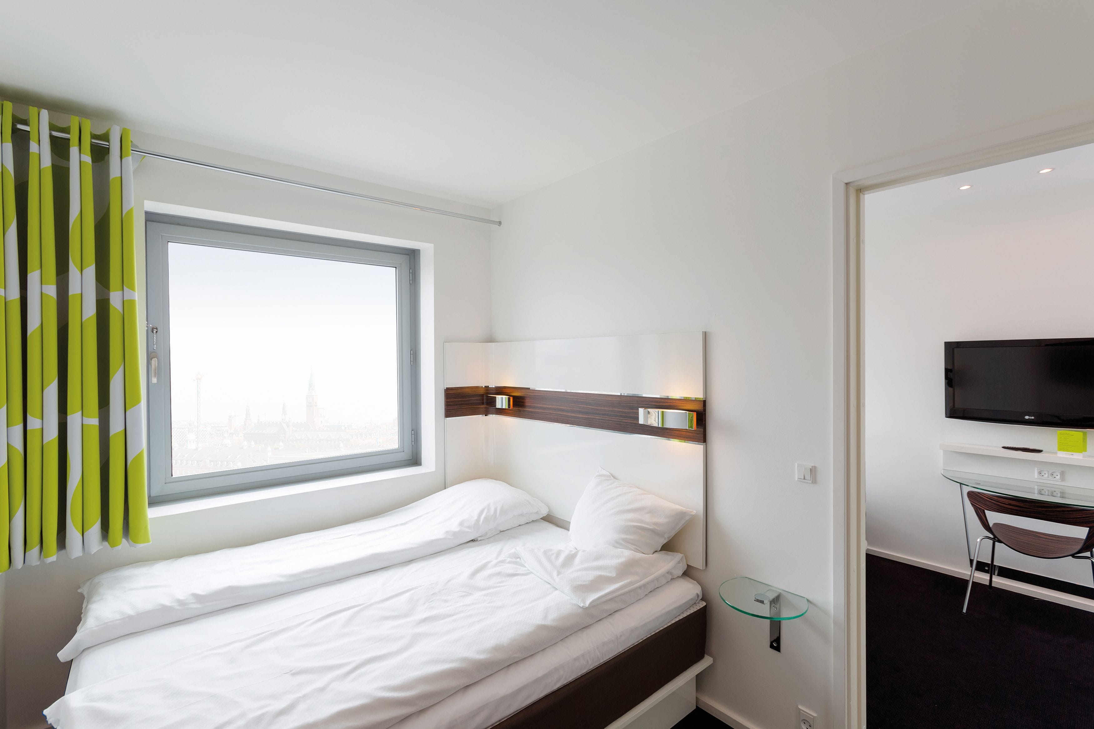 A hotel room with a bed, a TV, a desk, and a window with curtains.