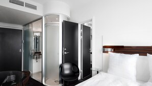 Une chambre d’hôtel moderne avec un lit, une douche en verre, une chaise noire et une petite table.