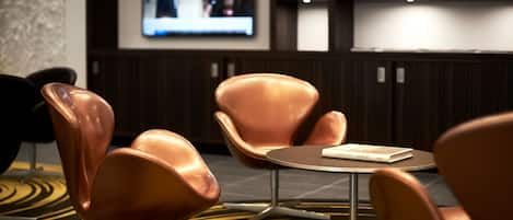 A modern hotel lobby with a round table, two cushioned chairs, and a television displaying a news broadcast.