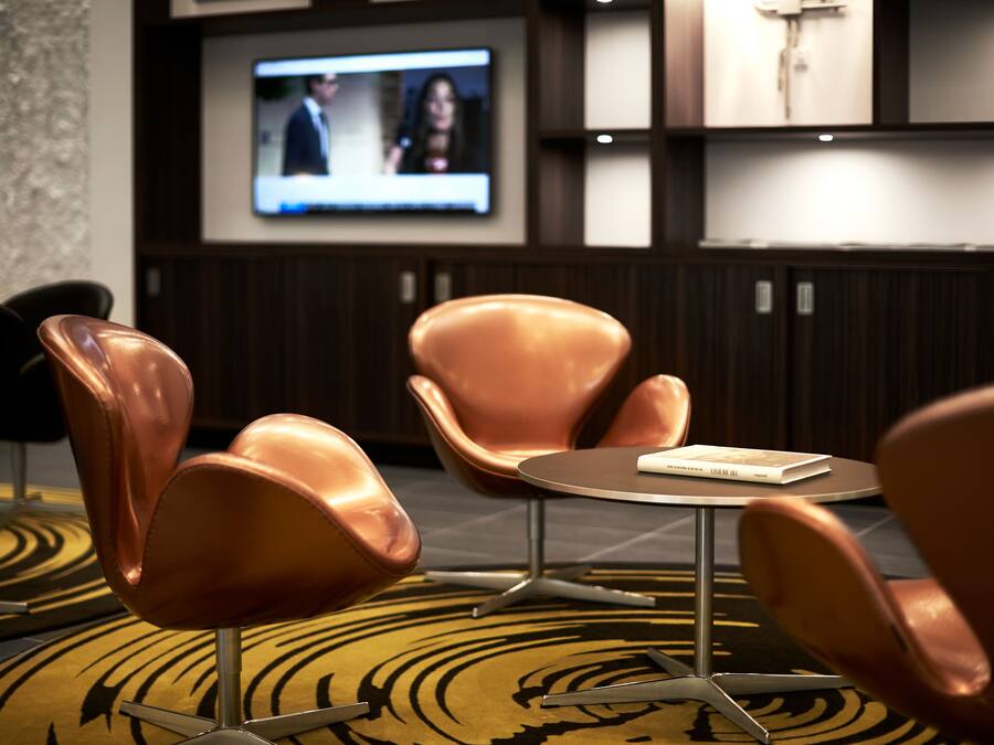Un'accogliente hall d'hotel moderna con un tavolino rotondo, due poltrone imbottite e un televisore che mostra un notiziario.