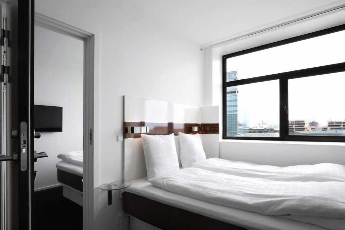 A modern hotel room with a large window, a bed with white linens, a bedside table, and a mounted TV.