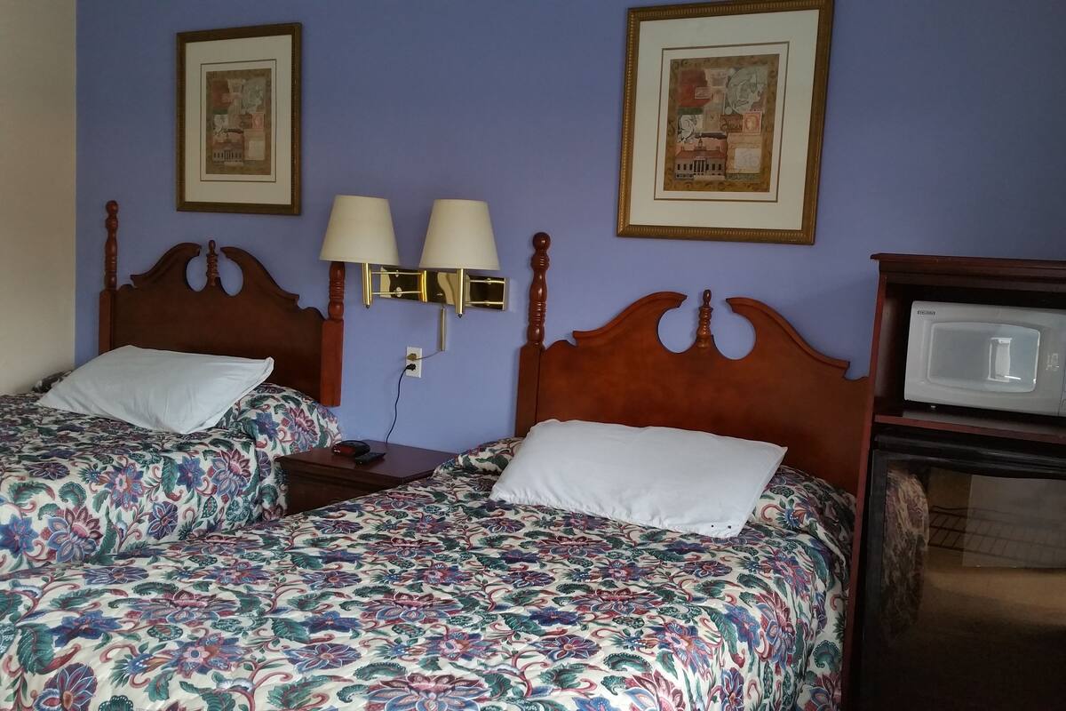 A hotel room with two beds, a nightstand, a wall-mounted lamp, a microwave, and two framed pictures on the wall.