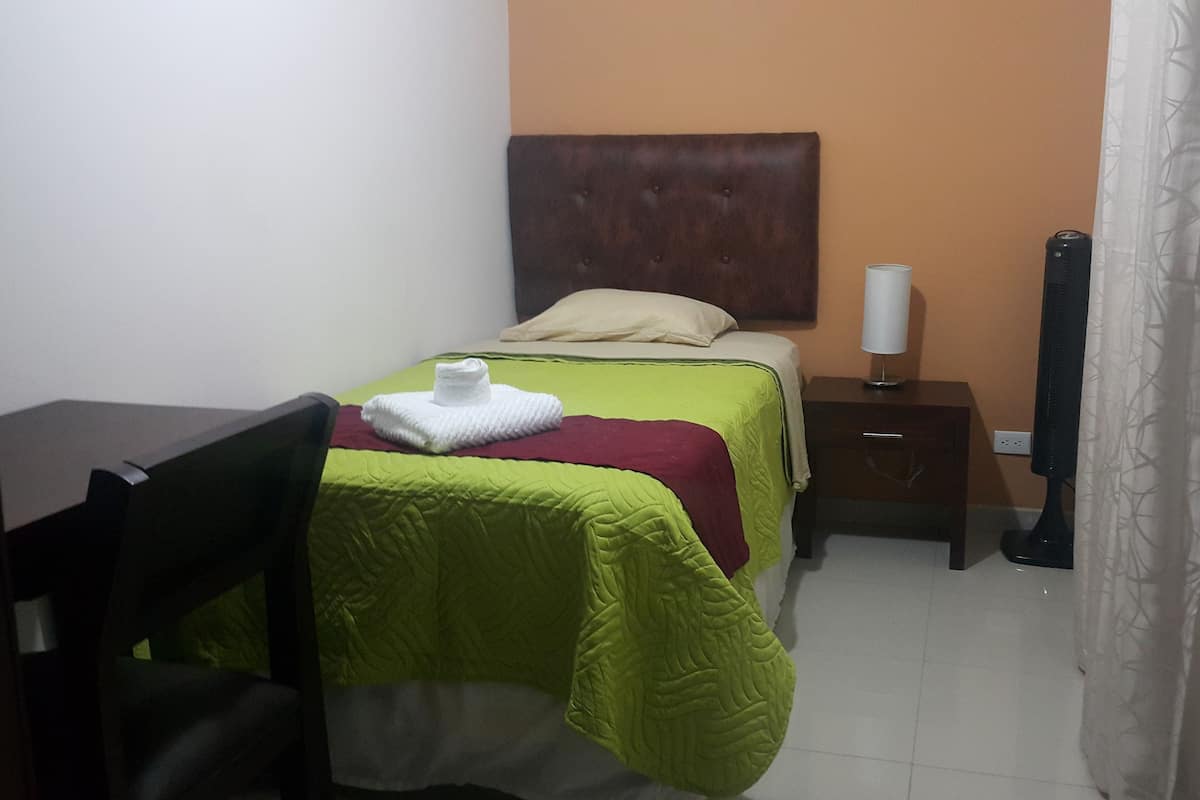 A small, neatly arranged bedroom with a bed, a bedside table, a lamp, and a chair.