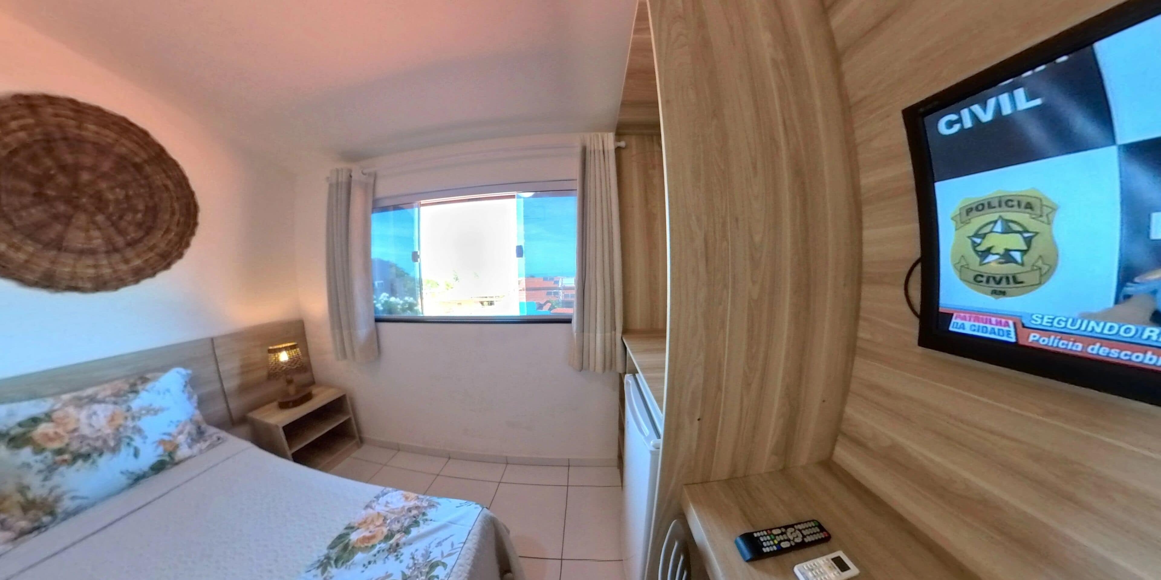 A hotel room with a bed, a bedside table, a TV screen displaying a news channel, and a window with curtains.