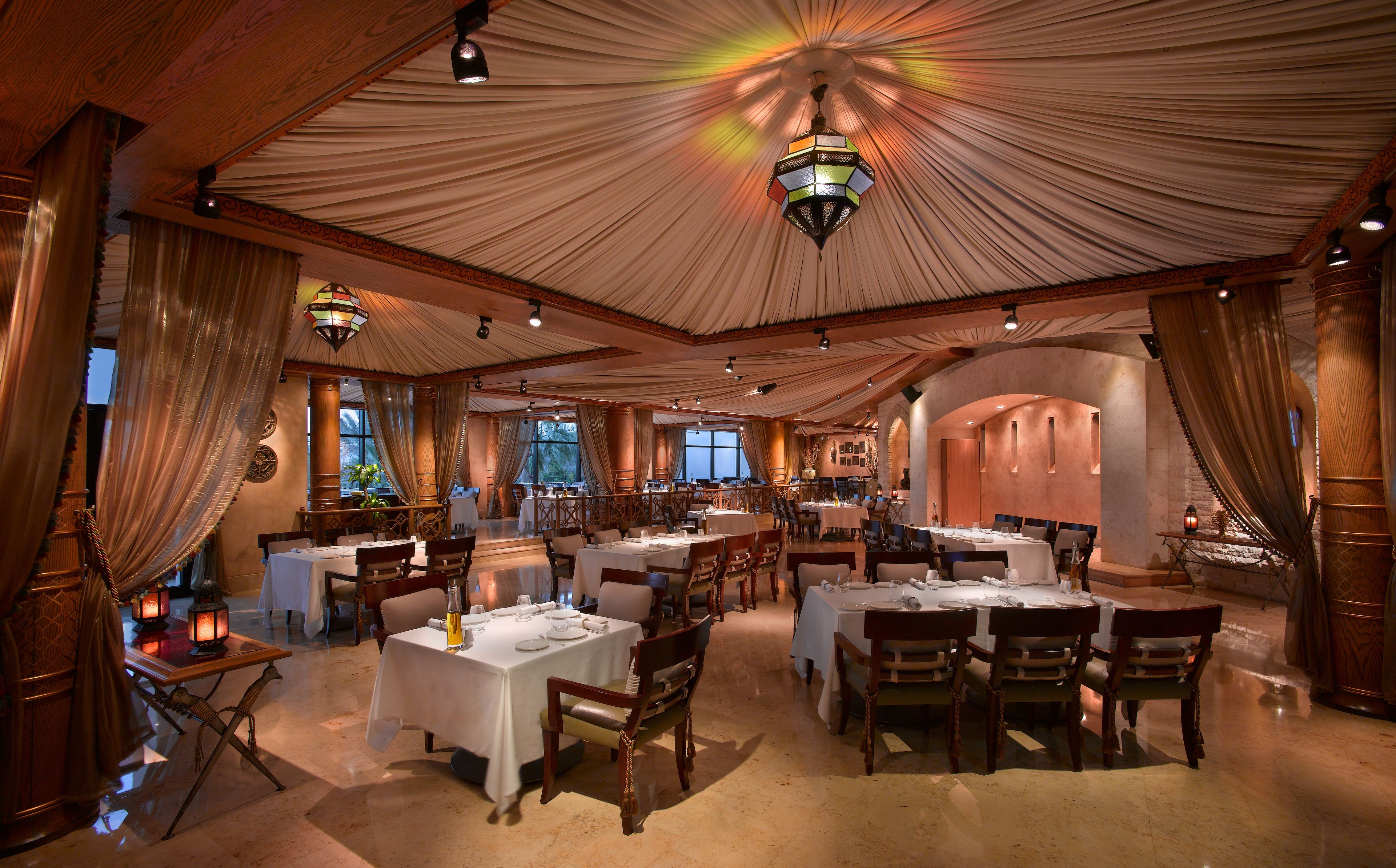 Ein geräumiges Restaurant mit mehreren Tischen und Stühlen, einer eleganten, drapierten Decke und warmer Beleuchtung.