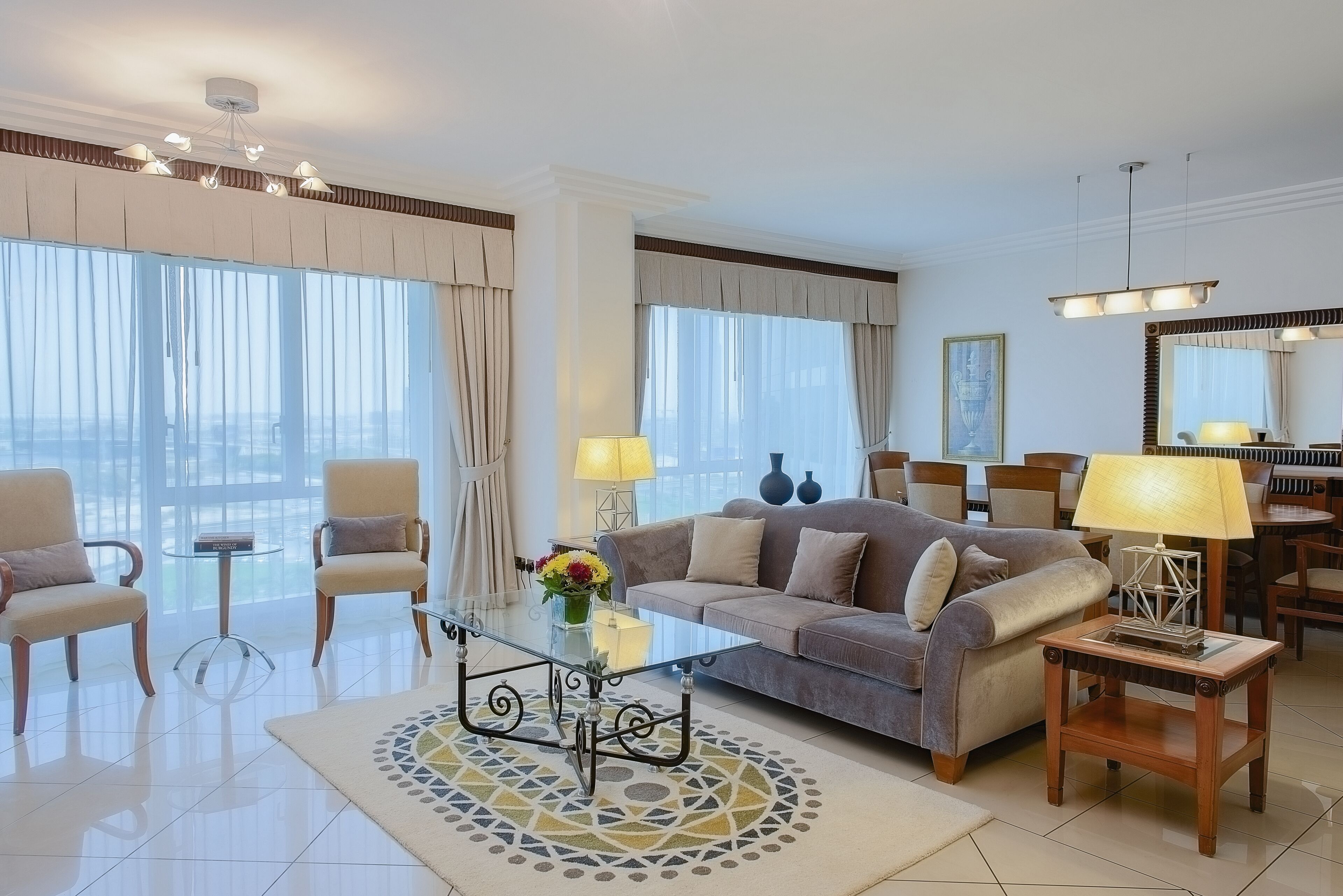 A modern living room with a glass coffee table, beige sofas, and a dining area with a chandelier.
