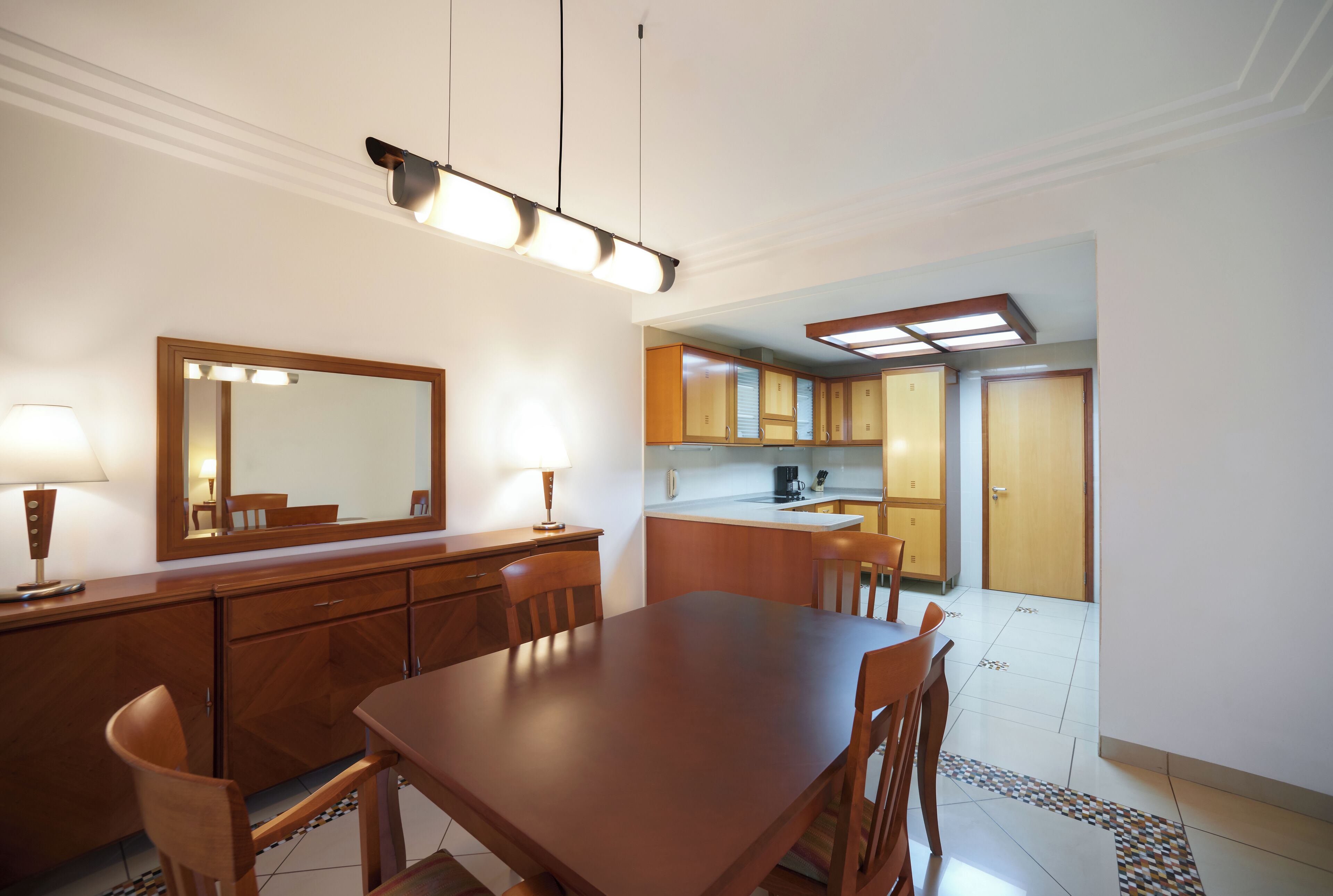 A dining area with a wooden table and chairs, a mirror, and a lamp. There is a kitchen in the background with wooden cabinets and a countertop.