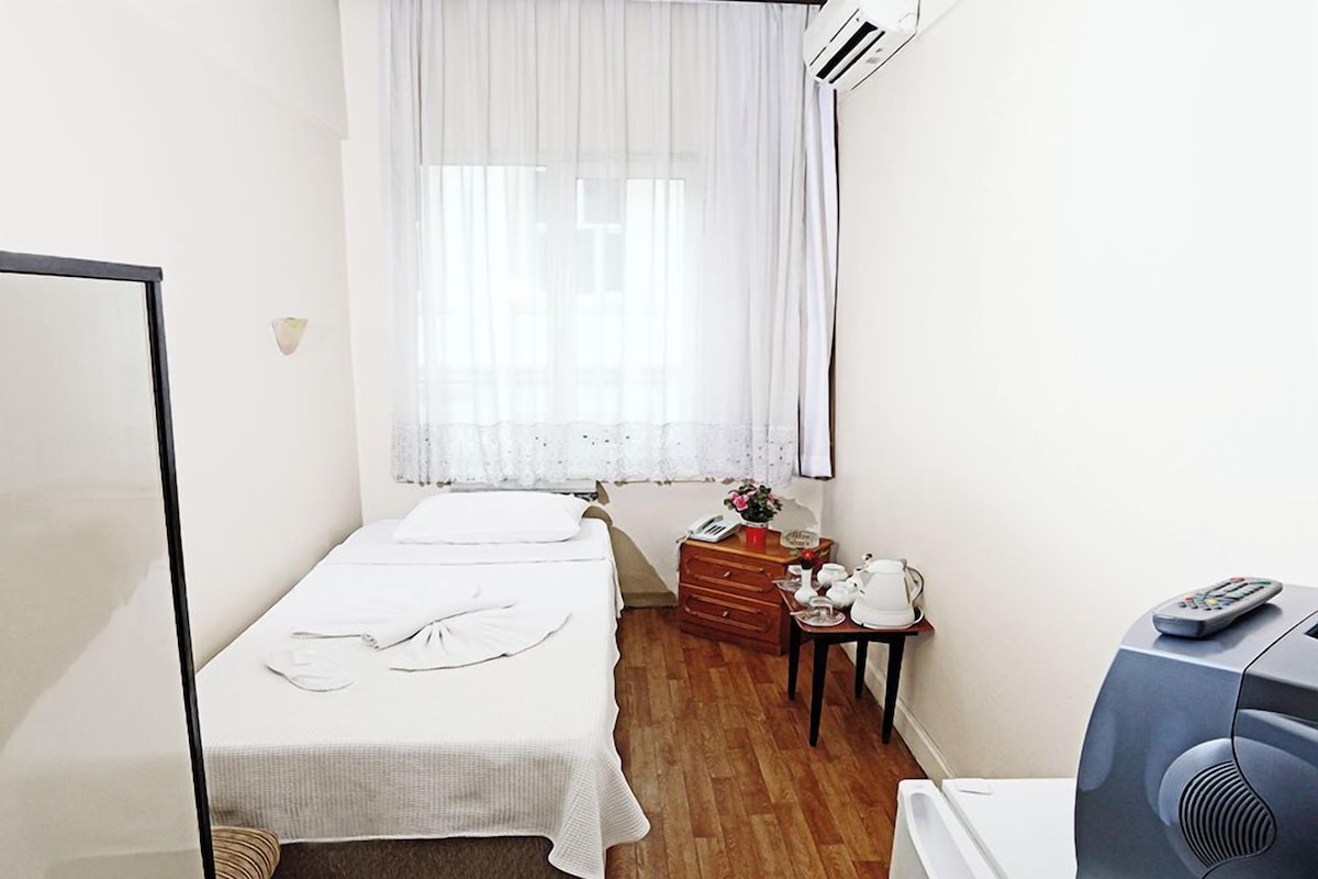 A small, clean room with two beds, a wooden dresser, a small table with a kettle, and a fan.