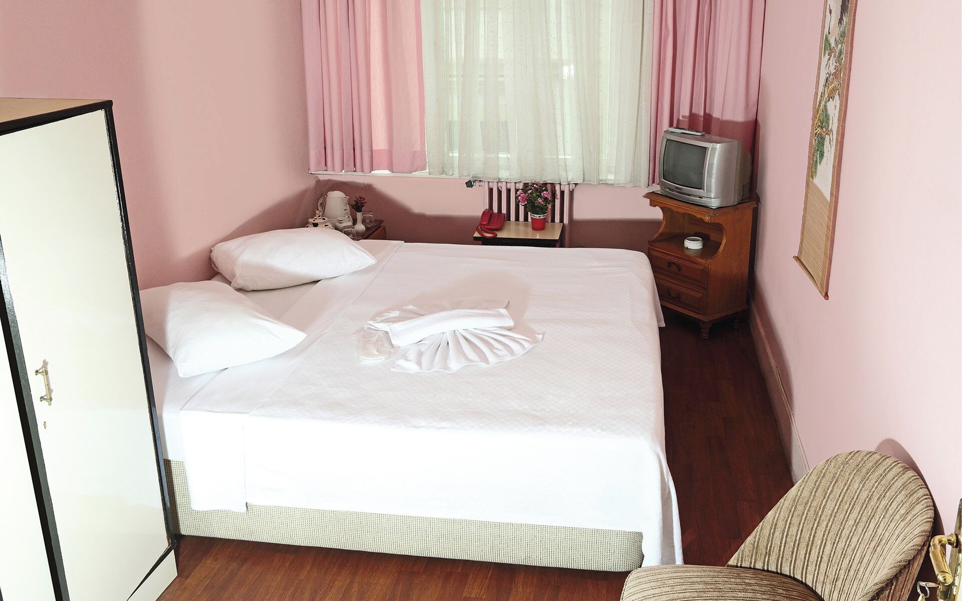 A neatly made bed with white linens, a bedside table with a lamp, a small TV, and a chair.