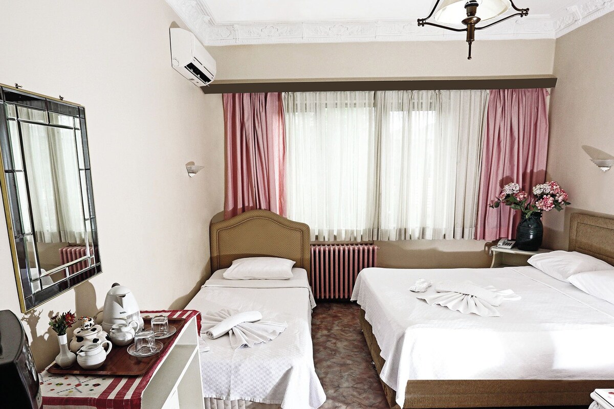 A hotel room with two beds, a mirror, a radiator, a table with a tray of items, and a vase with flowers.