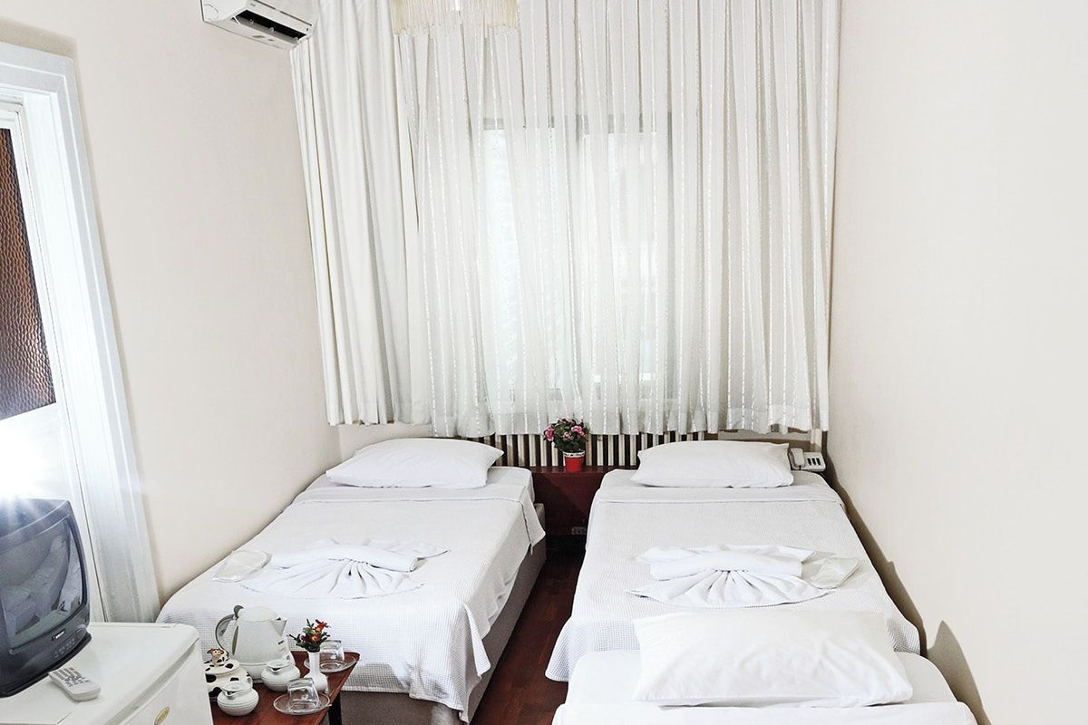 A hotel room with two beds, a TV, a small table with a pitcher and glasses, and a window with sheer curtains.
