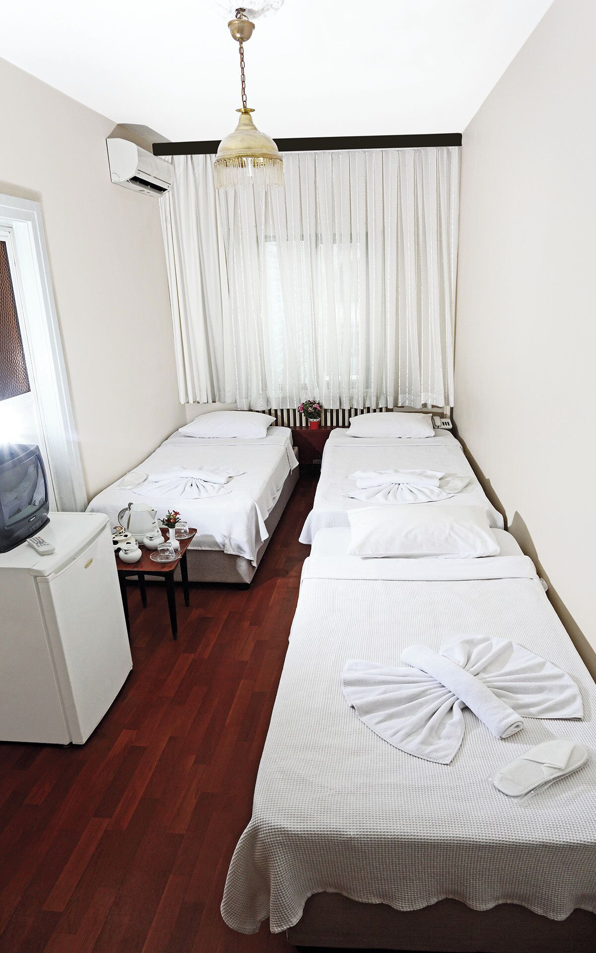 A hotel room with two beds, a small desk with a TV, a heater, and a window with sheer curtains.