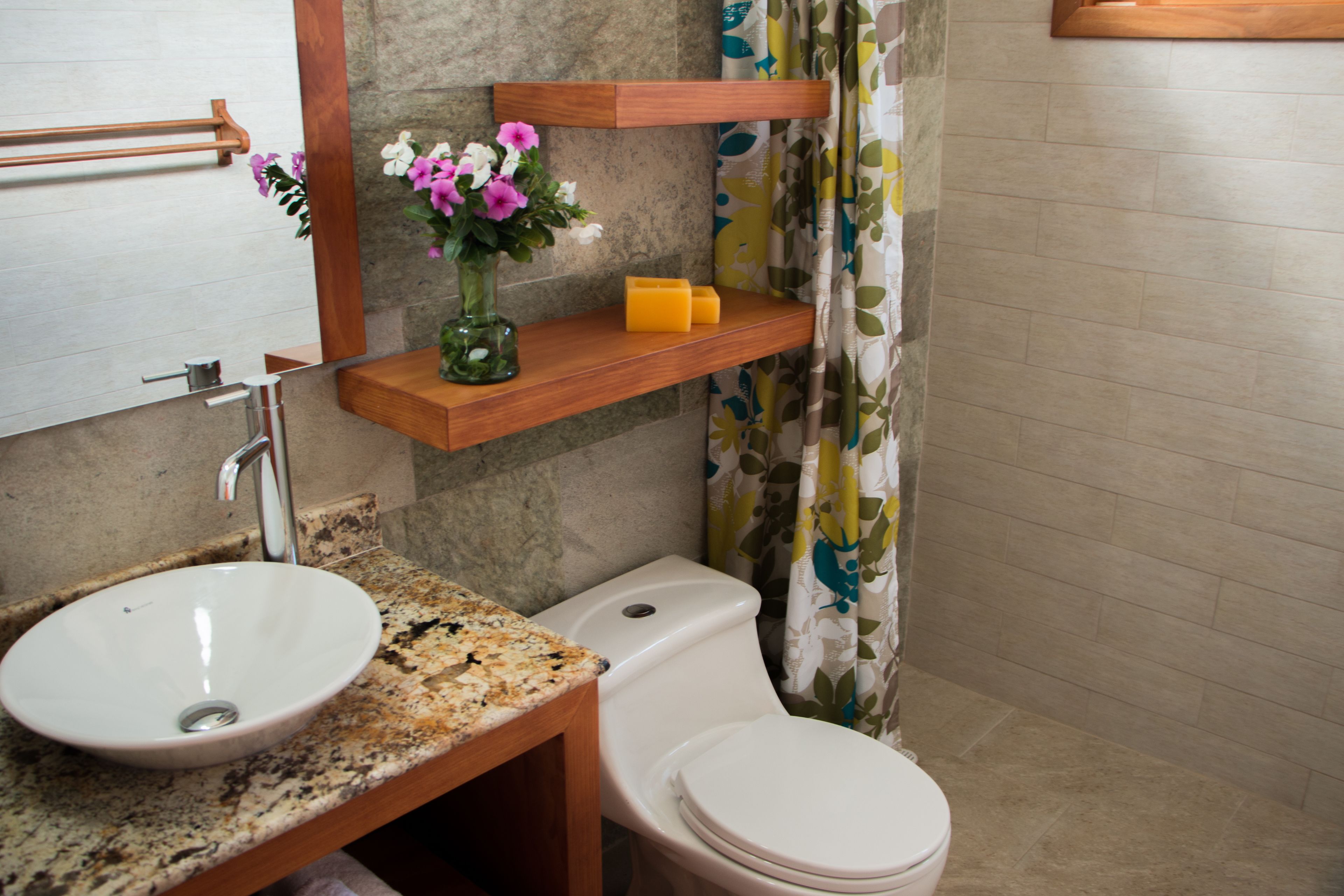 A bathroom with a granite countertop, white sink, and toilet. There is a wooden shelf above the toilet with a vase of flowers and a soap dish.