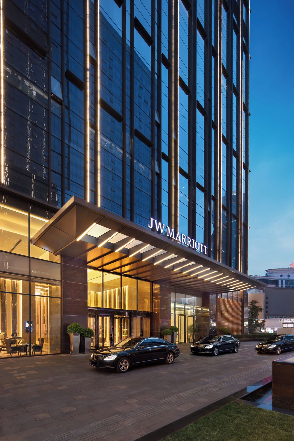 A JW Marriott hotel entrance with a modern glass facade, illuminated signage, and a parking area with several cars.