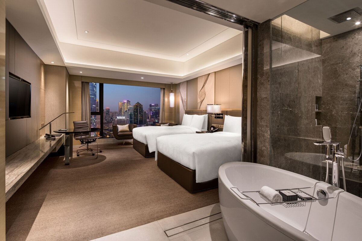 A modern hotel room with two beds, a large bathtub, a desk, and a city view.
