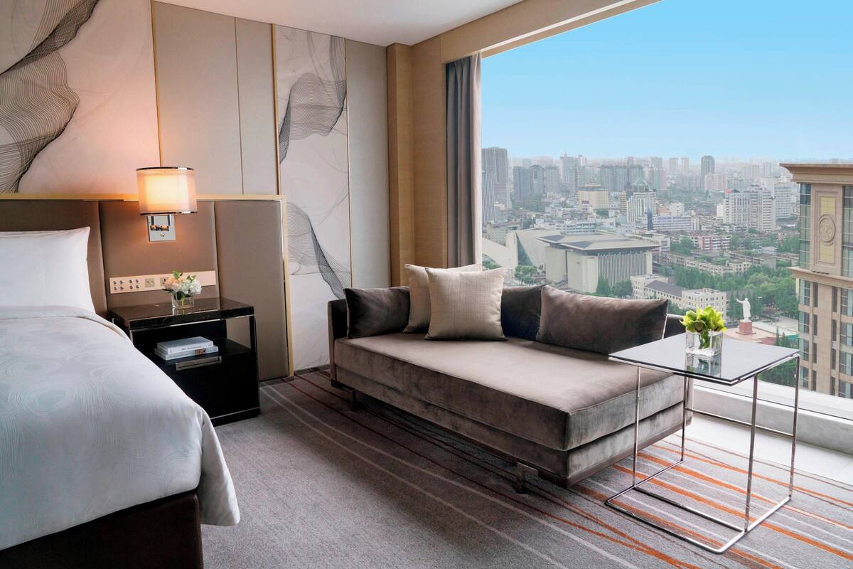 Una habitación de hotel con cama, sofá, mesita de noche con lámpara y vista a la ciudad.