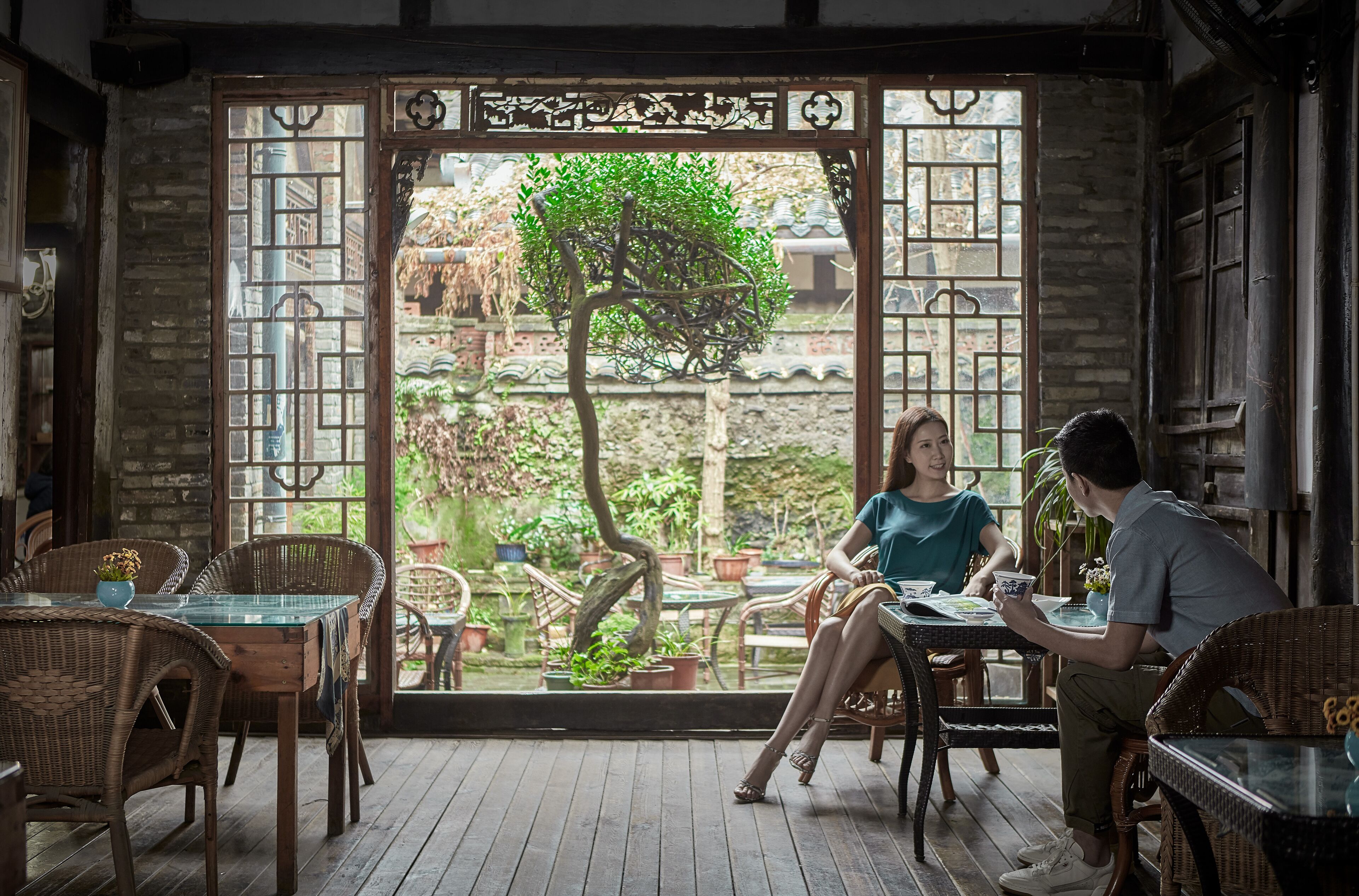 A traditional-style room with a wooden floor, wicker furniture, and a view of a garden through a decorative door.