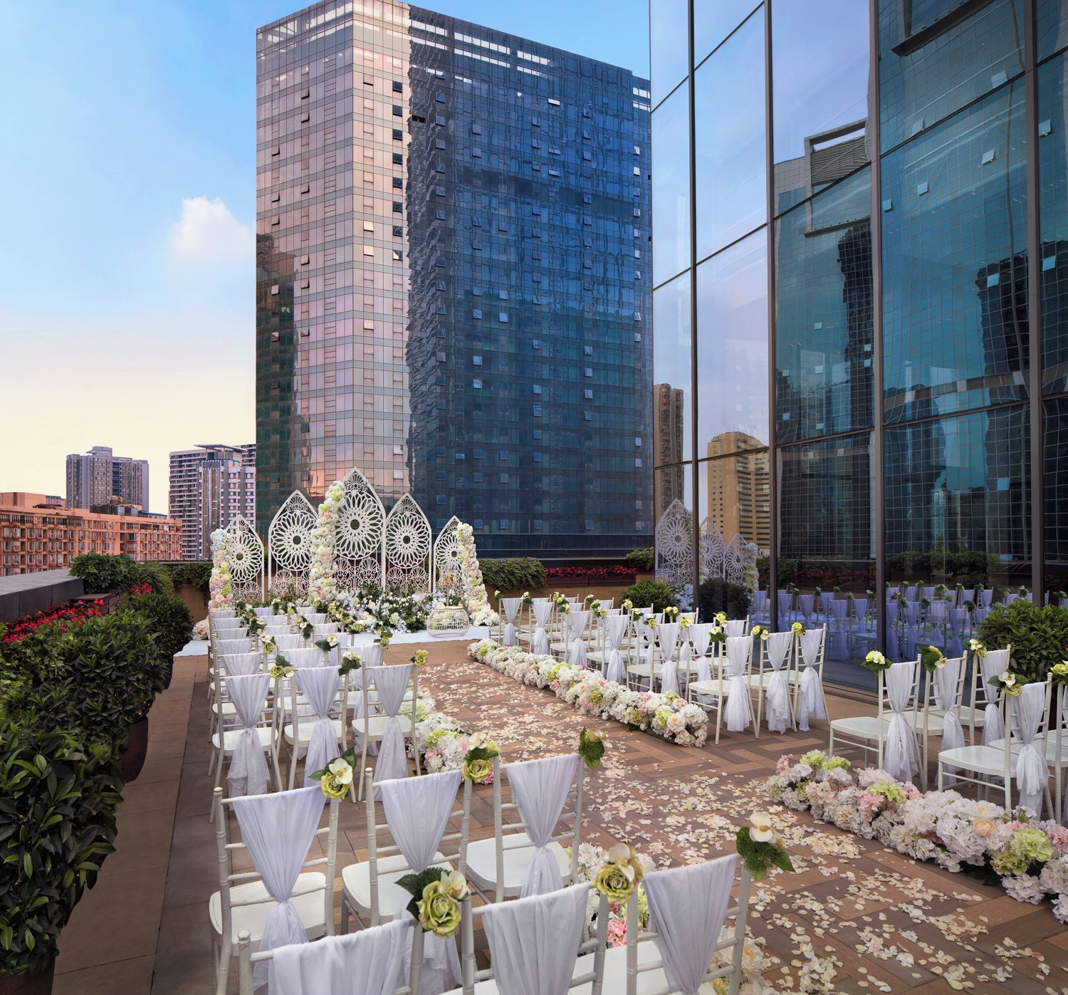 An outdoor wedding venue with white chairs and tables, floral decorations, and a cityscape in the background.