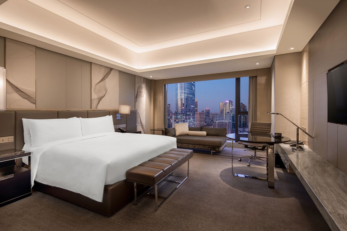 A modern hotel room with a large bed, a sofa, a desk, and a city view.