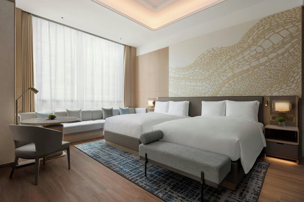 Habitación de hotel moderna con dos camas, un sofá, zona de comedor y un ventanal con cortinas translúcidas.