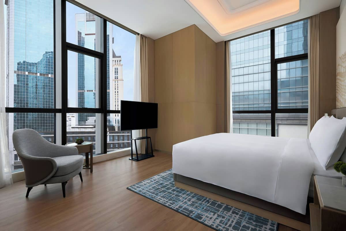 Habitación de hotel moderna con una cama grande, una silla, un televisor y vistas de la ciudad a través de amplios ventanales.