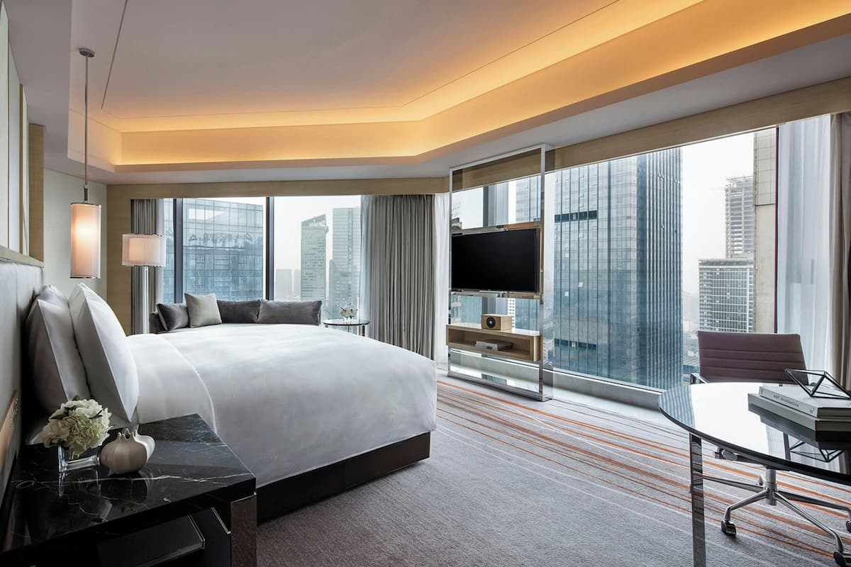 Habitación de hotel moderna con una cama grande, un escritorio, una silla, un televisor y un ventanal con vistas a la ciudad.