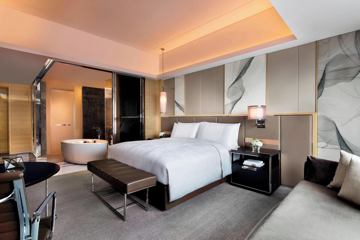 A modern hotel room with a large bed, a sitting area, a bathtub, and a desk with a lamp.