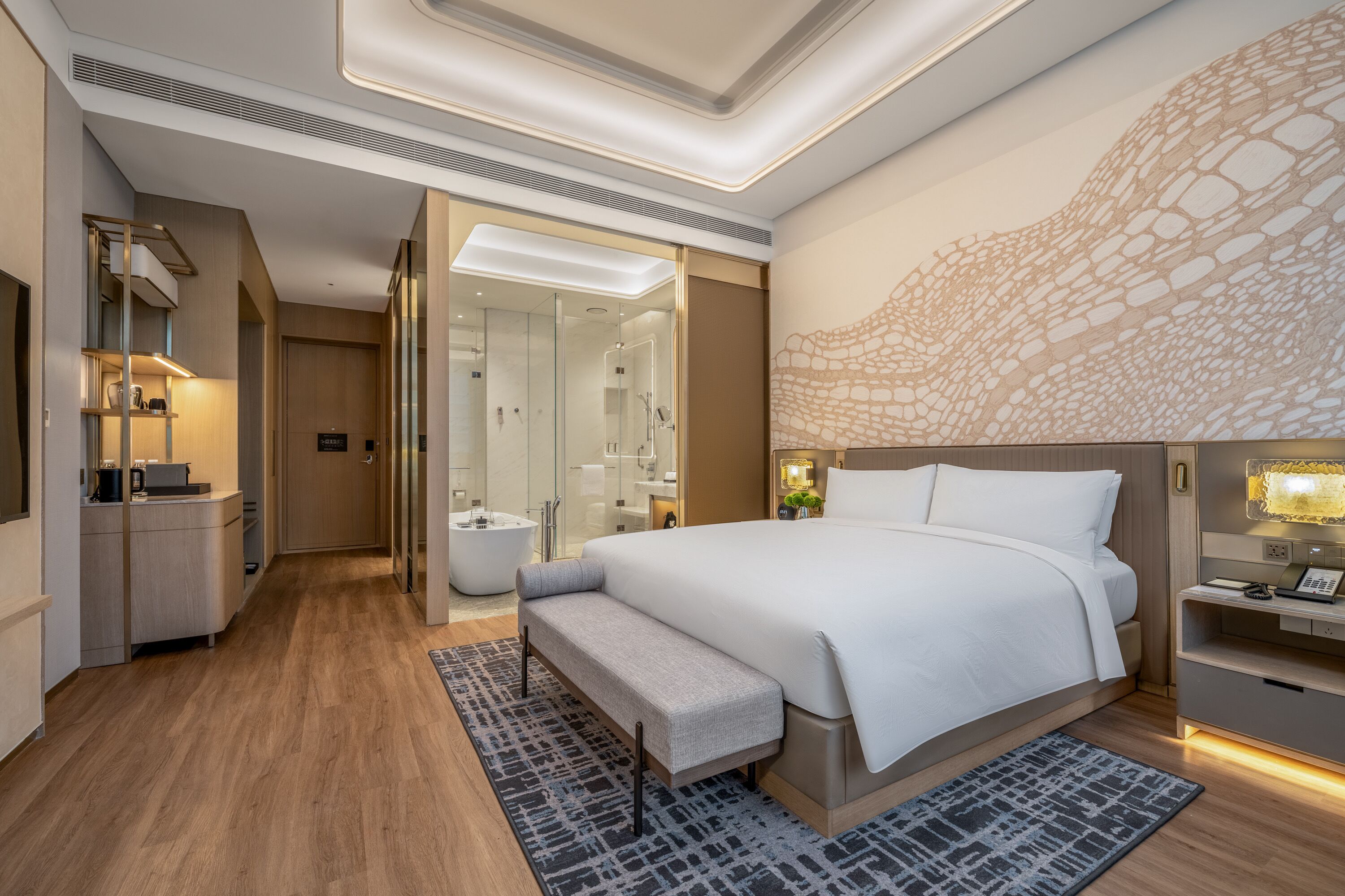 A modern hotel room with a large bed, a bench, a desk, and a bathroom with a glass shower.