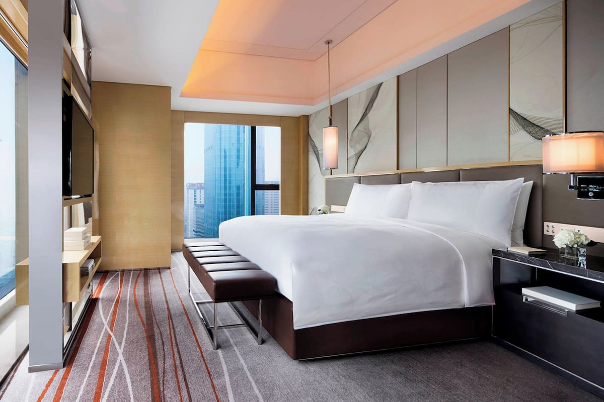 Habitación de hotel moderna con una cama grande, un banco, un televisor y vistas a la ciudad.