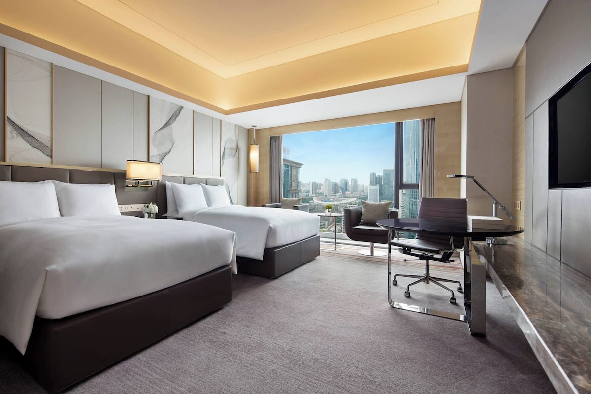 Habitación de hotel con dos camas, un escritorio y un amplio ventanal con vista a la ciudad.